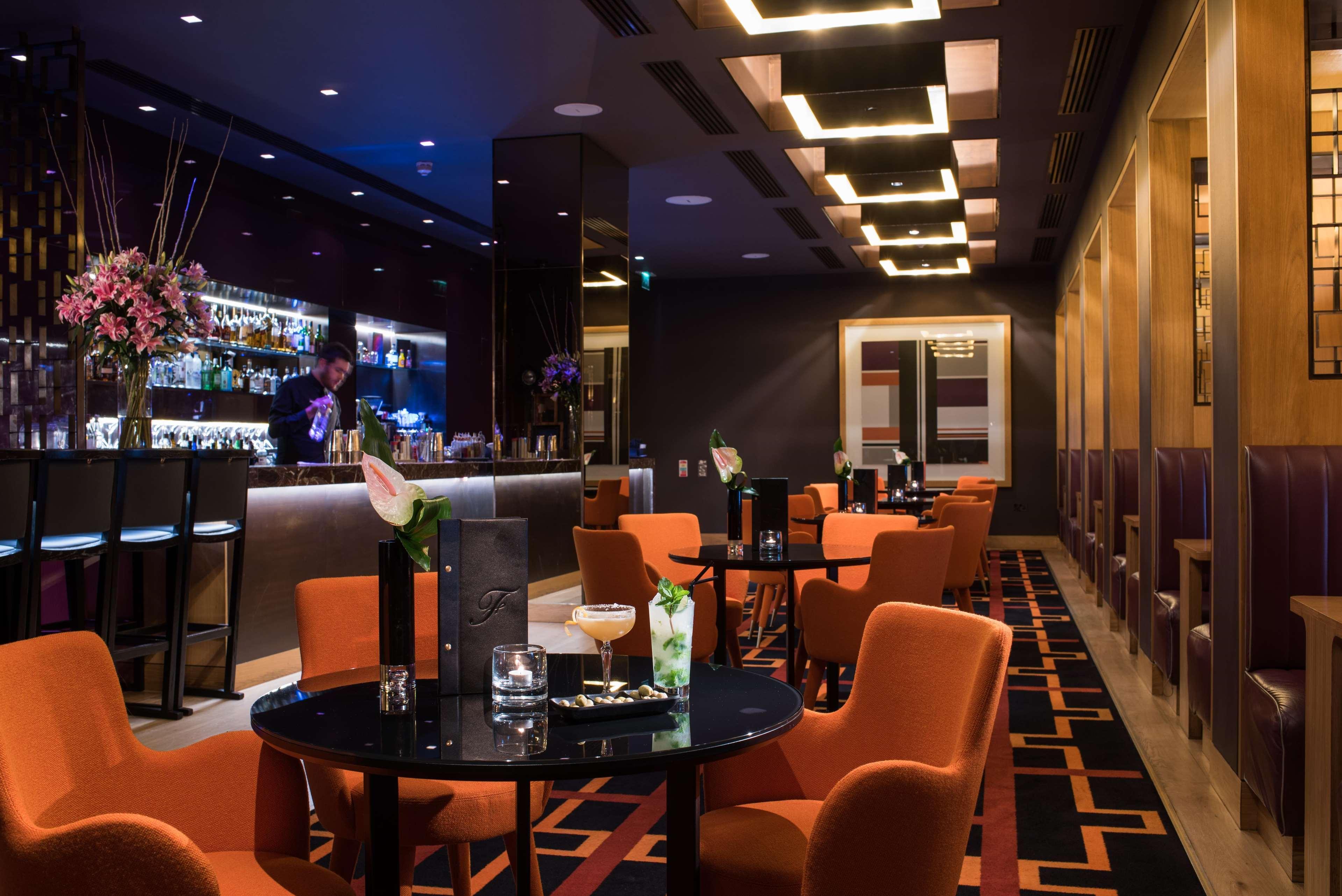 The Fitzwilliam Hotel Belfast Exteriör bild The photo depicts a stylish bar or lounge area with a modern and sophisticated atmosphere. The space features dark walls accented by blue lighting and a sleek bar counter in the background, stocked with various drinks. The seating consists of round t