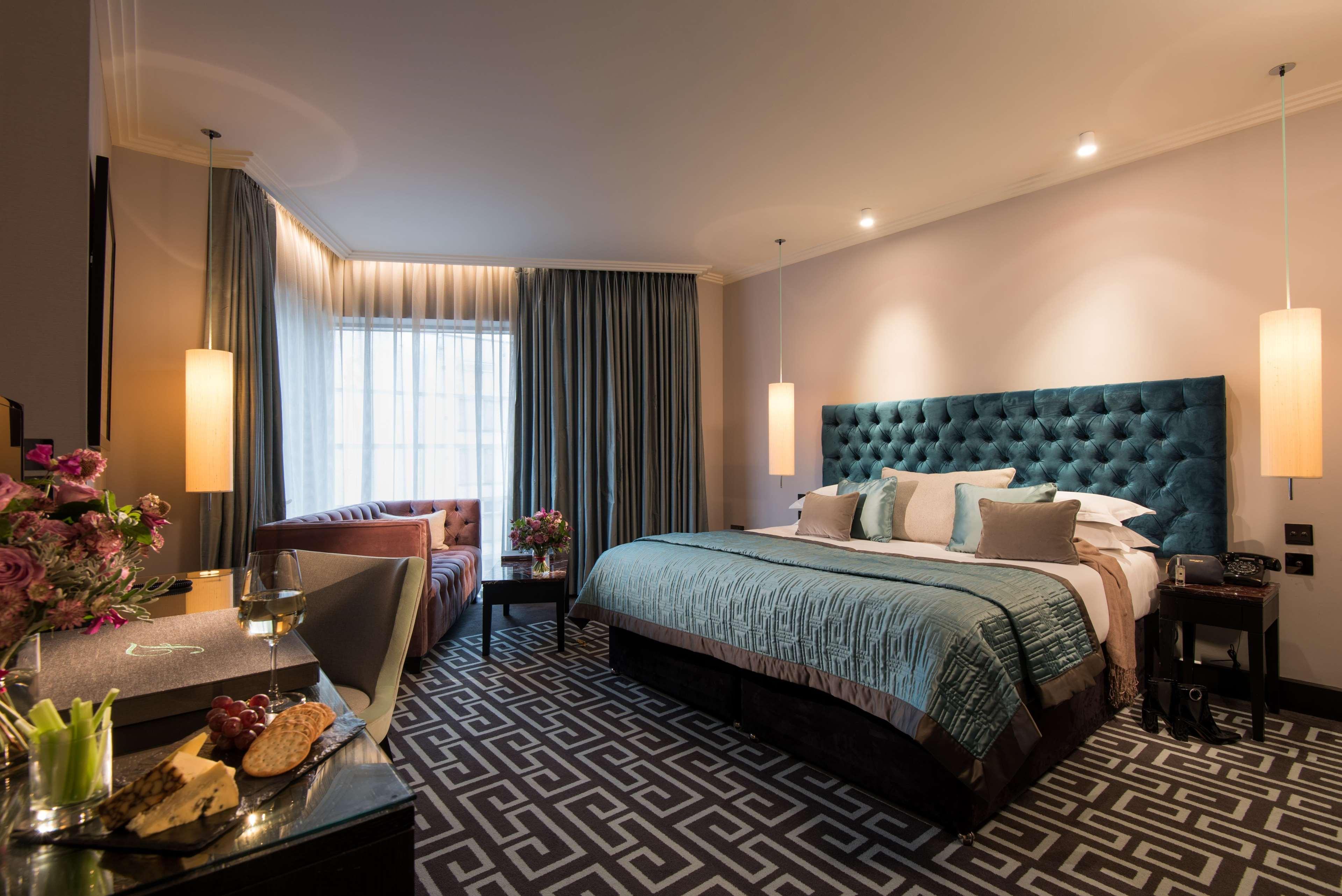 The Fitzwilliam Hotel Belfast Exteriör bild The photo depicts a stylish hotel room. It features a large bed with a tufted teal headboard, flanked by two wall sconces providing soft lighting. The bedding is neatly arranged with accent pillows. To one side, there is a small seating area with a c