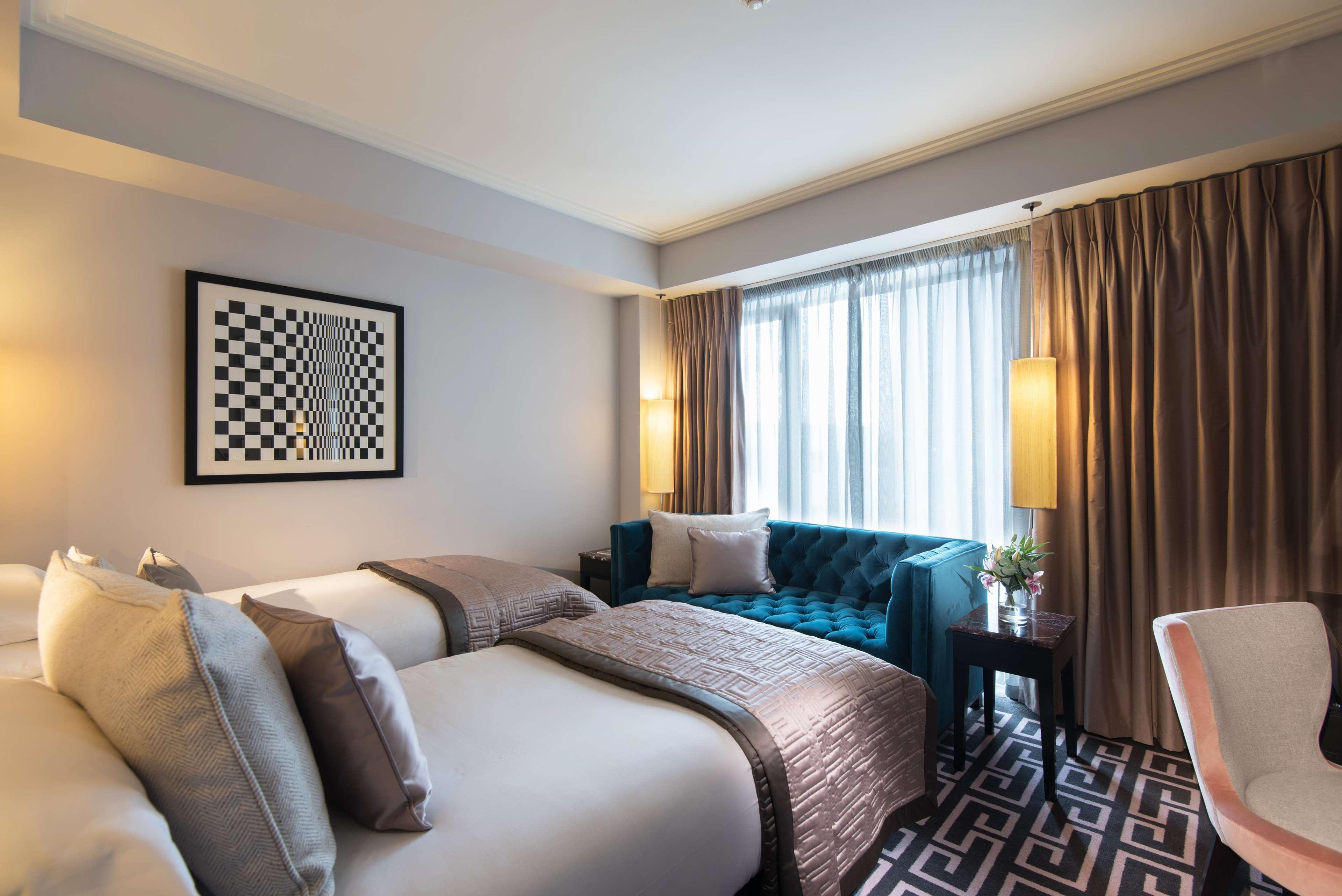 The Fitzwilliam Hotel Belfast Exteriör bild The photo shows a modern hotel room. It features two beds with decorative pillows, a stylish teal couch, and large windows with sheer curtains allowing natural light to enter. The walls are painted in neutral tones, and there is a piece of artwork wi