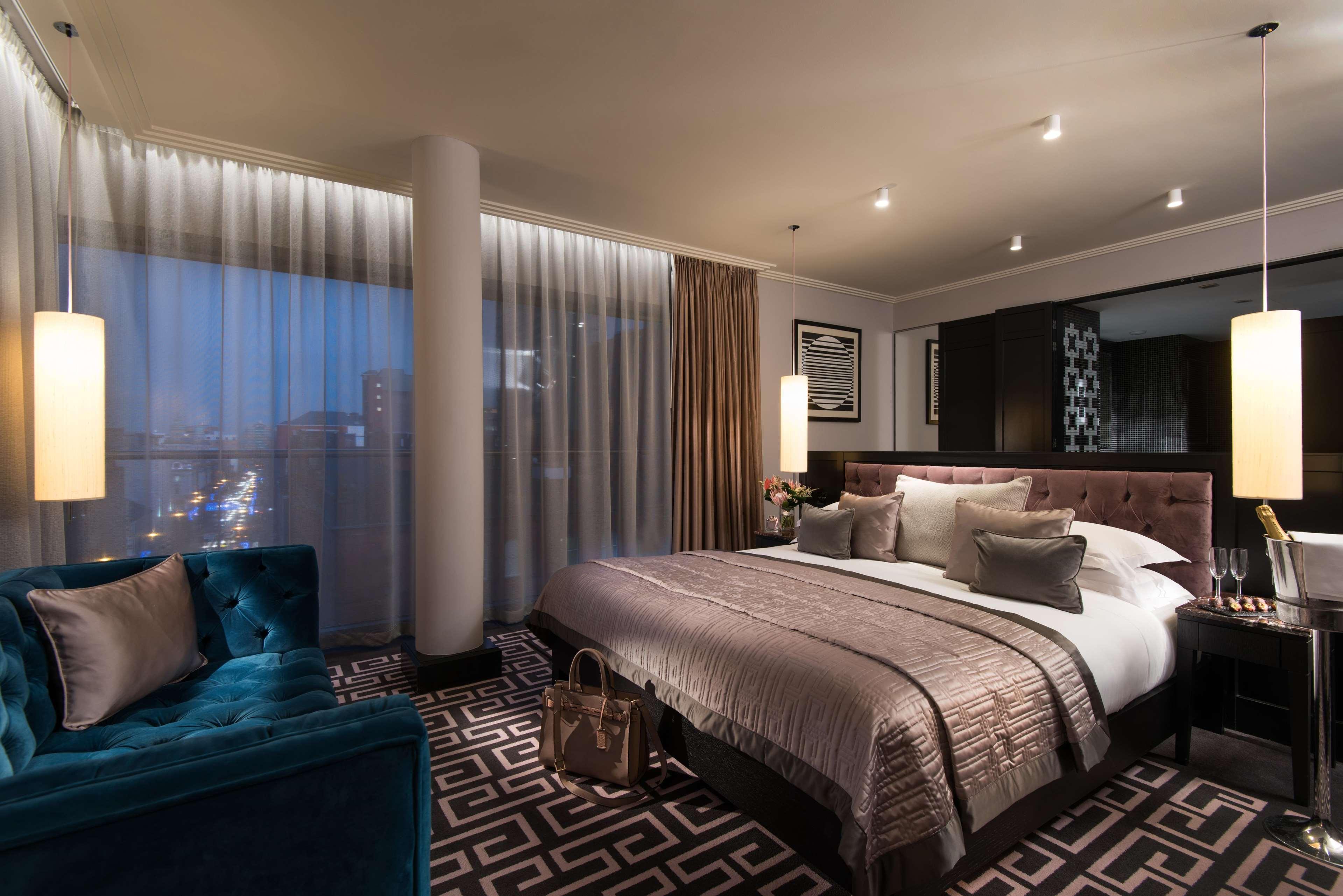 The Fitzwilliam Hotel Belfast Exteriör bild The photo shows a luxurious hotel room with a modern design. It features a large bed with decorative pillows and a stylish bedspread, complemented by bedside lamps. To one side, there is a plush turquoise sofa and a coffee table. The room has floor-t