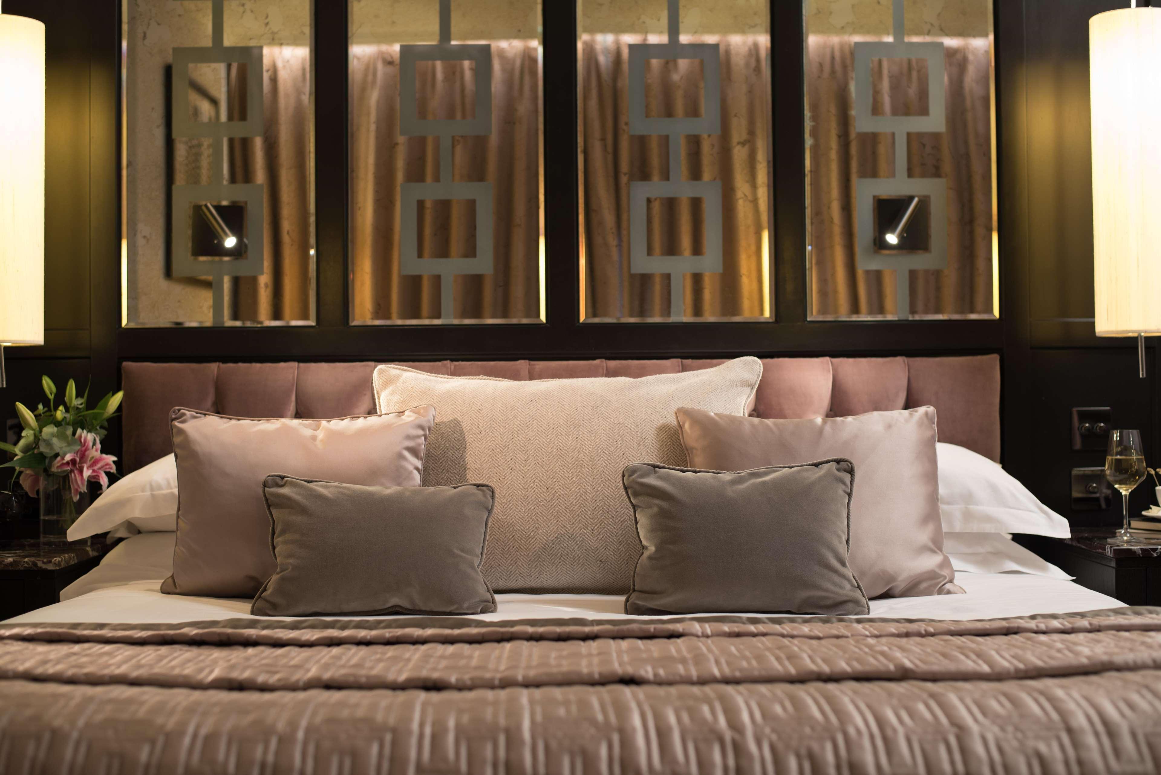 The Fitzwilliam Hotel Belfast Exteriör bild The photo shows a beautifully designed bed in a stylish bedroom. The bed is adorned with a soft, plush headboard and layered with various decorative pillows in shades of beige and gray. The bedding appears luxurious, with a textured blanket. Behind t