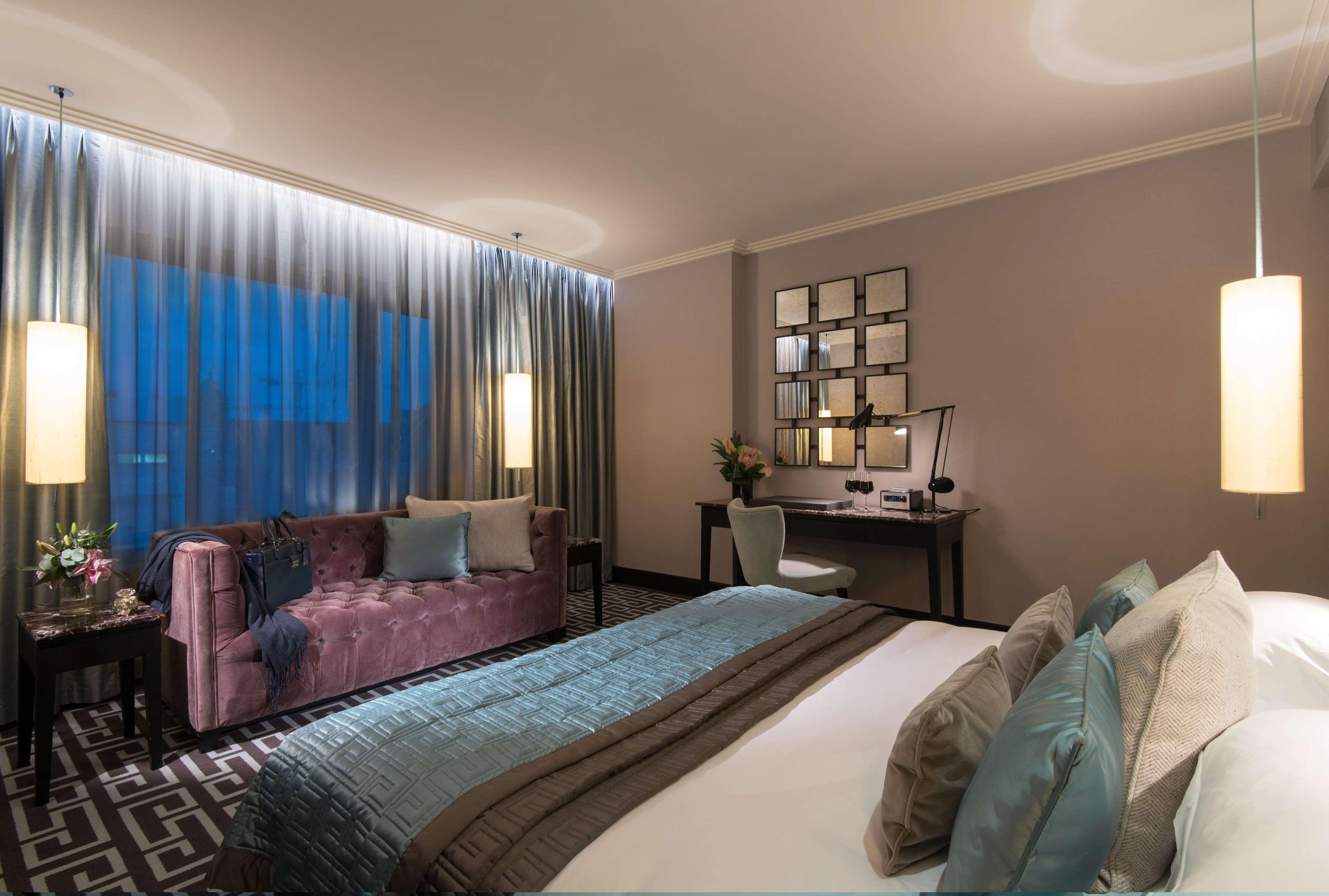 The Fitzwilliam Hotel Belfast Exteriör bild The photo depicts a modern hotel room featuring a spacious layout. There is a large bed with decorative pillows in the foreground, covered with a stylish bedspread. To one side, a plush purple sofa adds a pop of color and comfort. A small desk and ch
