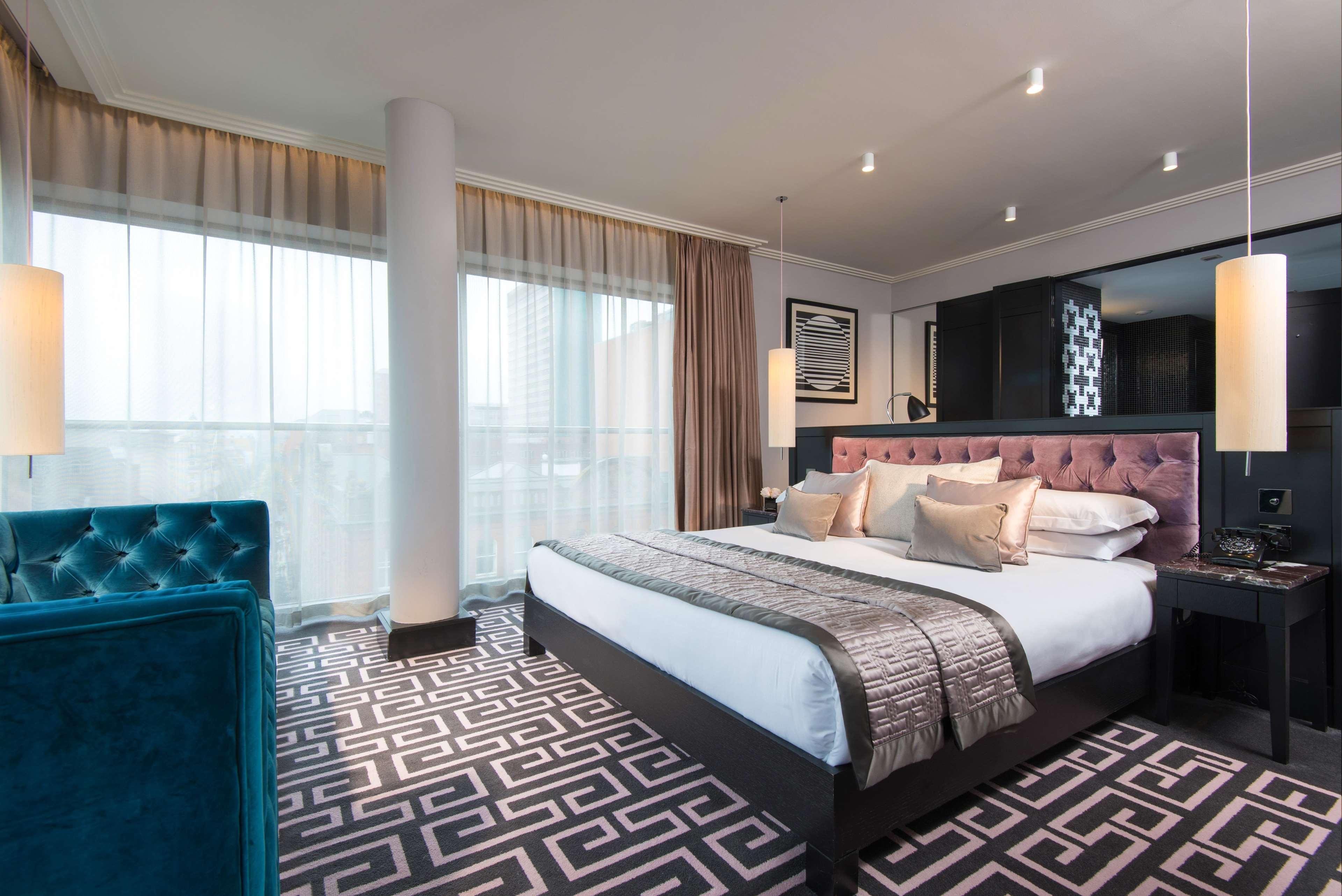 The Fitzwilliam Hotel Belfast Exteriör bild The photo showcases a modern hotel room. It features a large bed with plush pillows and a stylish headboard. The room has large windows with sheer curtains, allowing natural light to filter in. The floor is covered with a patterned carpet that contra